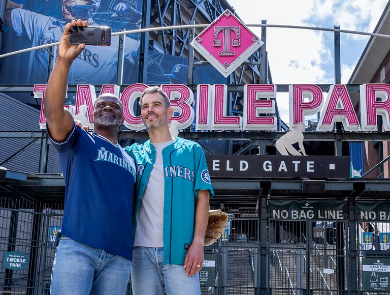 Dos amigos posan para un selfie a la entrada de un campo de béisbol.