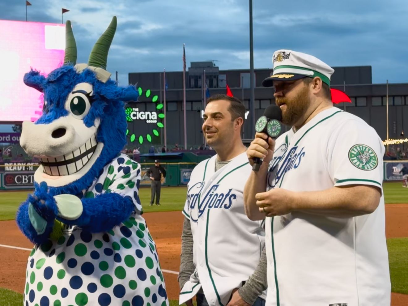 Detective Rafael Dacampo, a Hartford police detective, stands on a baseball field beside a team mascot and another person.