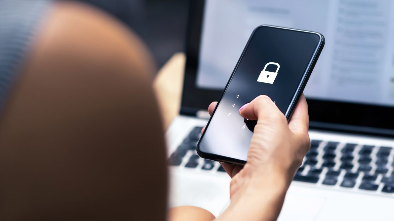 Primer plano de unas manos que sostienen un smarphone​​​​​​​ con un ícono de candado superpuesto y una laptop en el fondo.