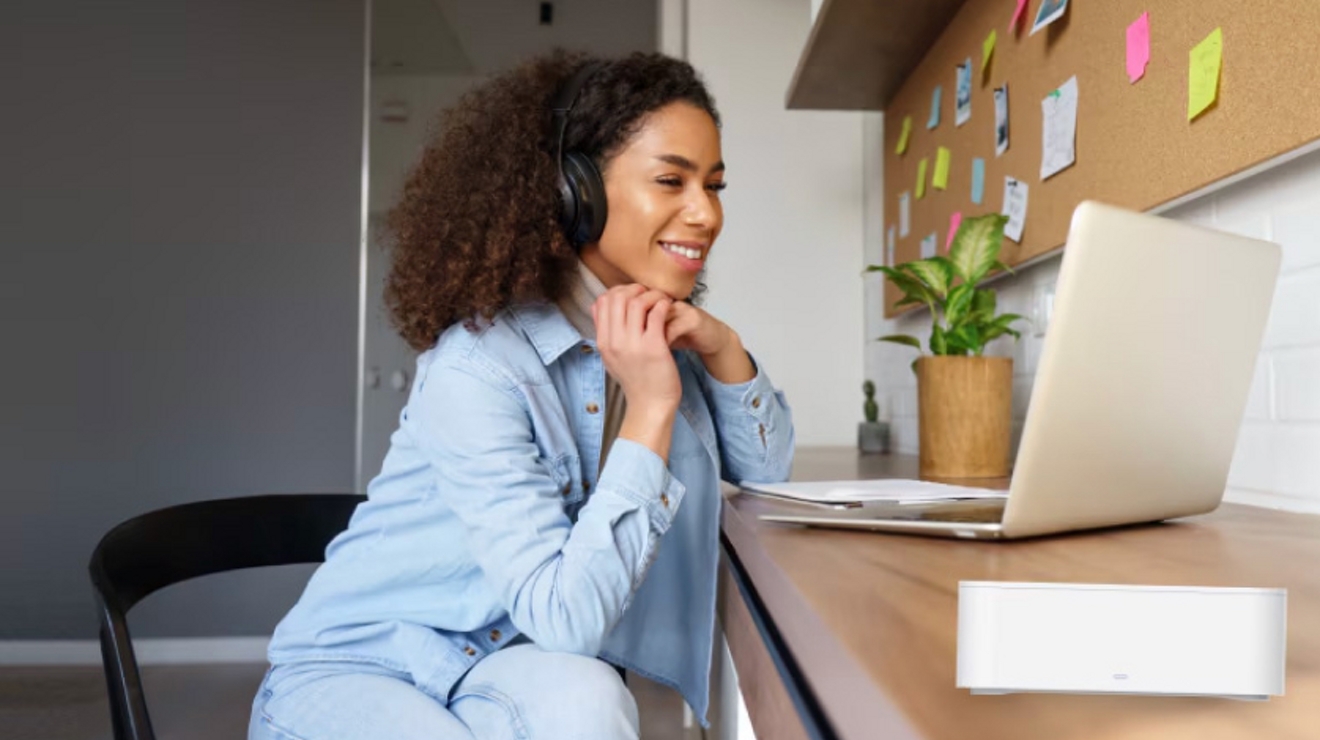 Estudiante con audífonos sentada a su escritorio sonríe a la pantalla de su laptop.