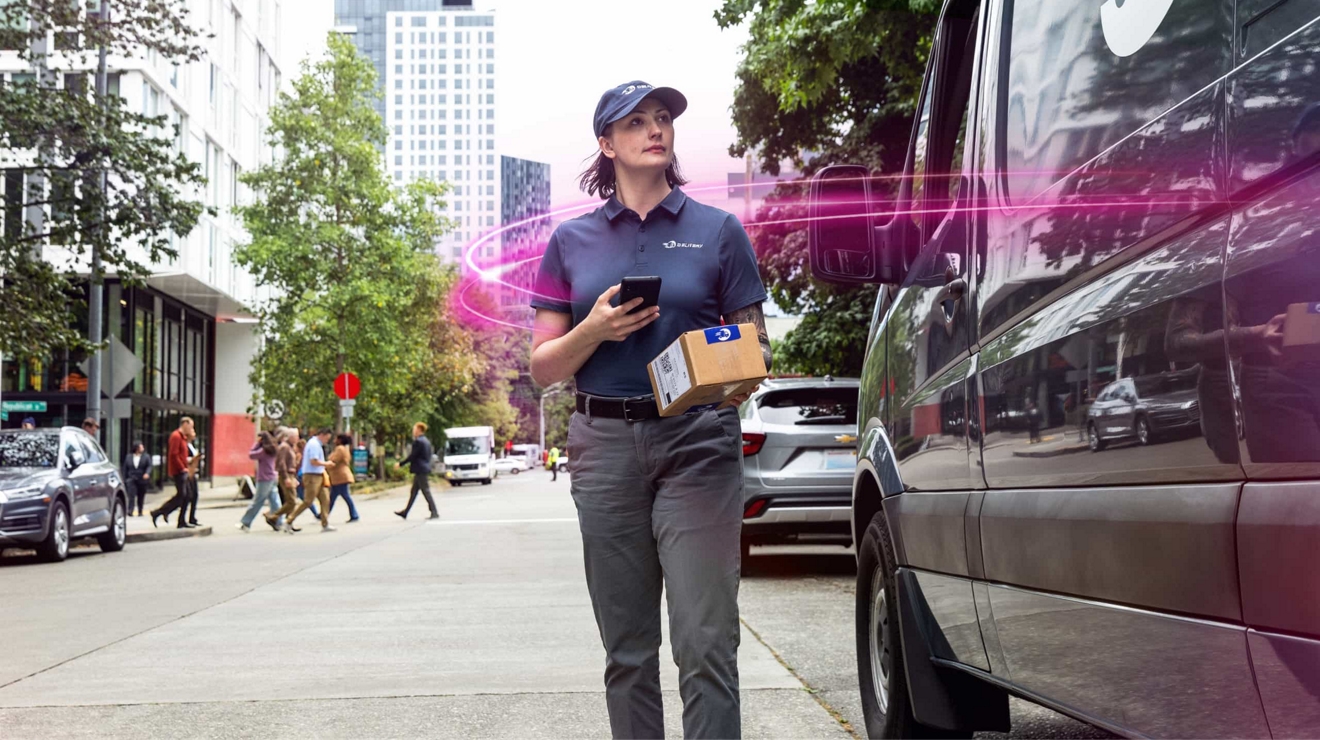 Una mujer con uniforme de reparto camina por una acera de la ciudad sosteniendo un paquete y un teléfono, mientras unos rayos de luz magenta la rodean mientras está al lado de una camioneta.