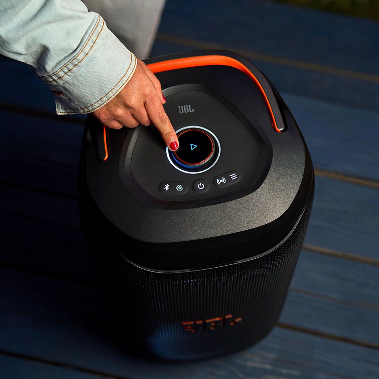 Una mano presiona el botón de reproducción iluminado en la parte superior de una bocina JBL PartyBox 130 negra con iluminación de decorativa naranja.