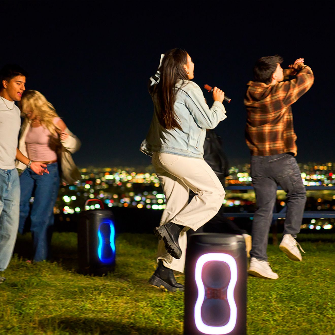 Dos bocinas JBL PartyBox 130 con luces LED de colores reproducen música mientras un grupo de amigos baila y canta con un micrófono al aire libre por la noche, con vistas al horizonte de la ciudad.