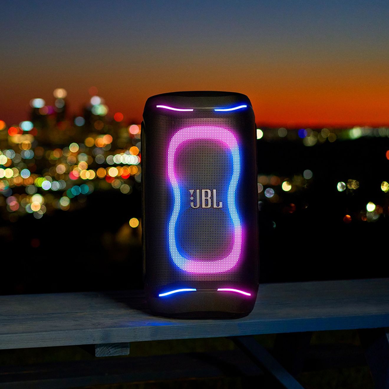 Una bocina JBL PartyBox 130 negra muestra un colorido espectáculo de luces LED azules y rosas sobre un horizonte urbano borroso por la noche.