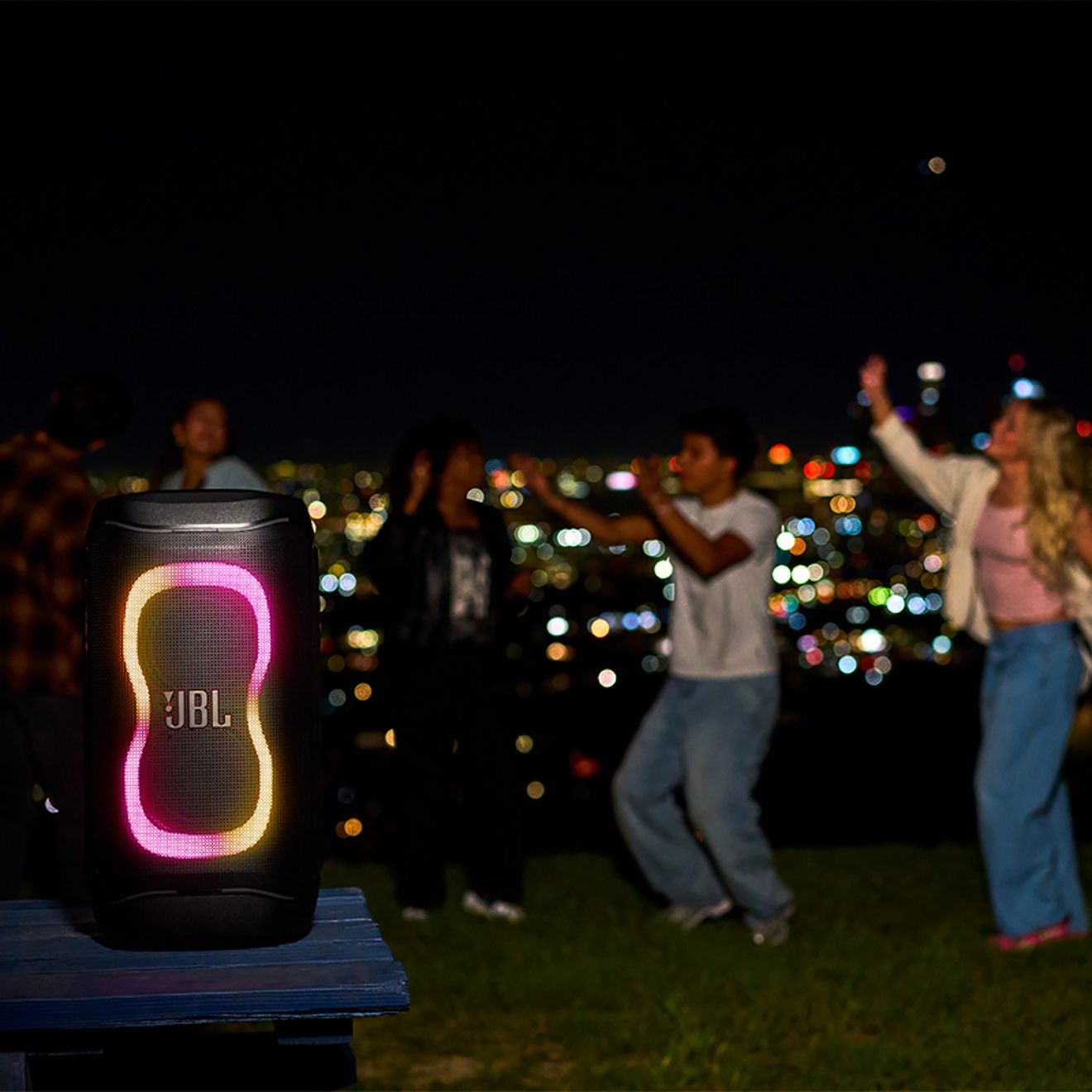 Una bocina JBL PartyBox 130 con luces LED de colores suena en una fiesta nocturna al aire libre mientras un grupo de amigos baila con el horizonte de la ciudad iluminado de fondo.