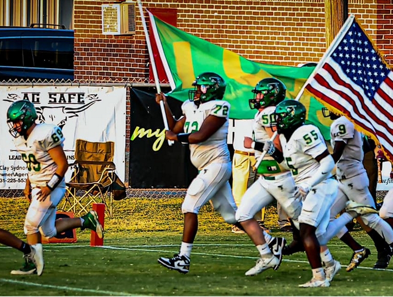 El equipo de fútbol de Lake County High School, con uniformes en blanco y verde, corre al campo de juego llevando banderas.