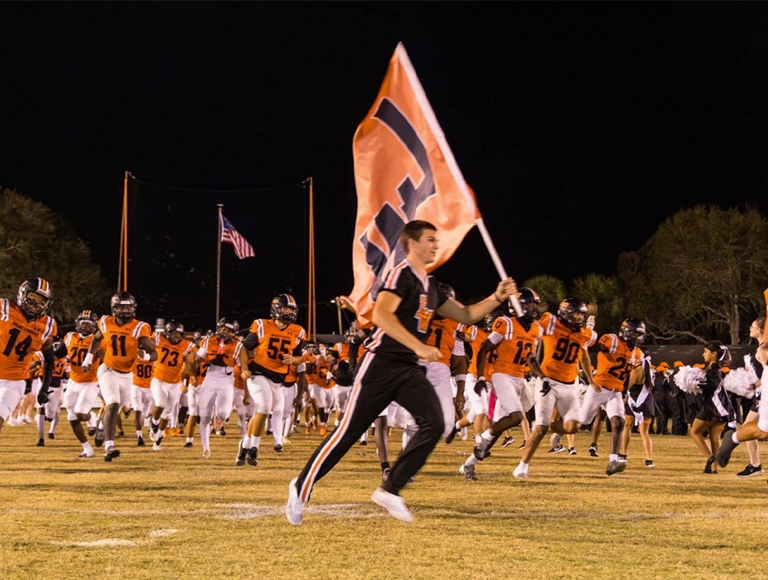 El equipo de fútbol de Lake Wales, con uniformes en naranja y negro, corre al campo mientras una porrista ondea una bandera de Lake Wales.