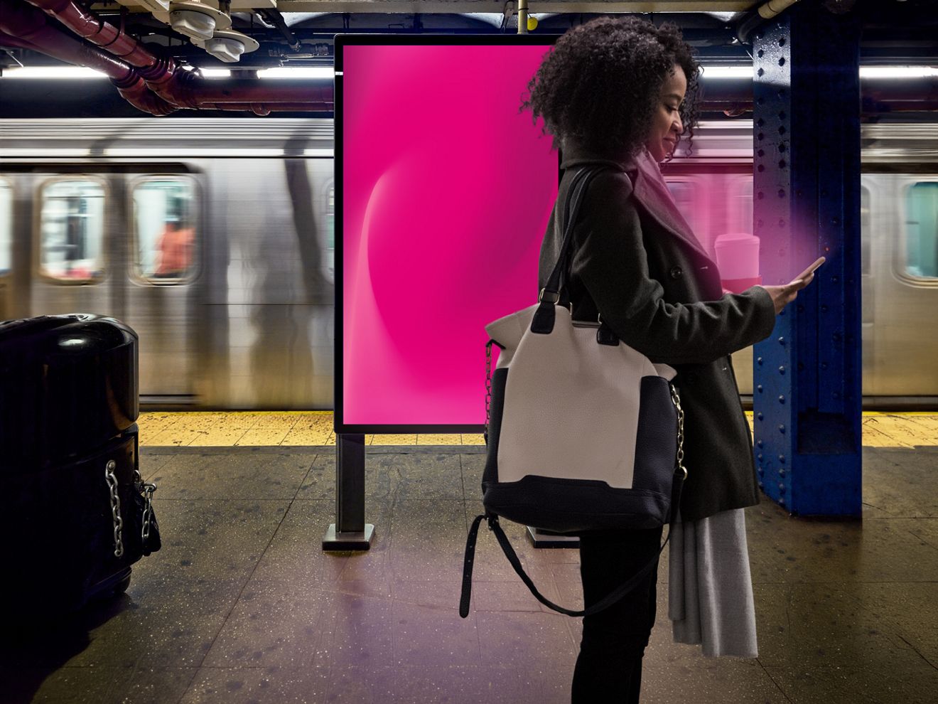 Una mujer usando el teléfono cerca de una pantalla digital exterior, con un tren de transporte público detrás de ella.