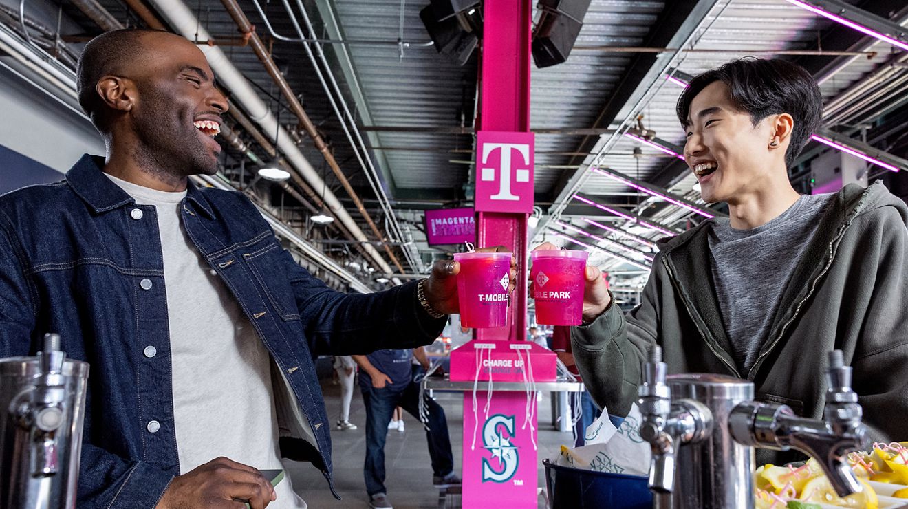 Two Seahawks fans toasting with Magenta-colored drinks at a concession stand inside T-Mobile Park.