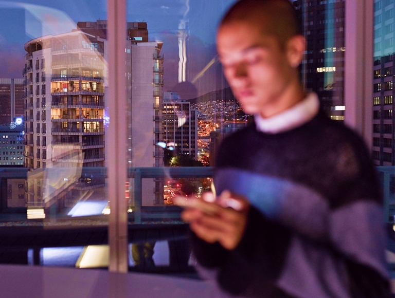 Una persona está de pie junto a la ventana de su apartamento en la ciudad y revisa los mensajes de su teléfono.