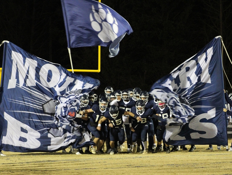 El equipo de fútbol americano de Mount Airy, con uniformes azul marino y blanco, corre por una pancarta hacia el campo de juego.