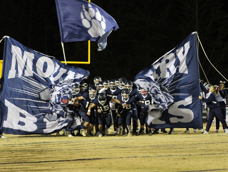 El equipo de fútbol americano de Mount Airy, con uniformes azul marino y blanco, corre por una pancarta hacia el campo de juego.