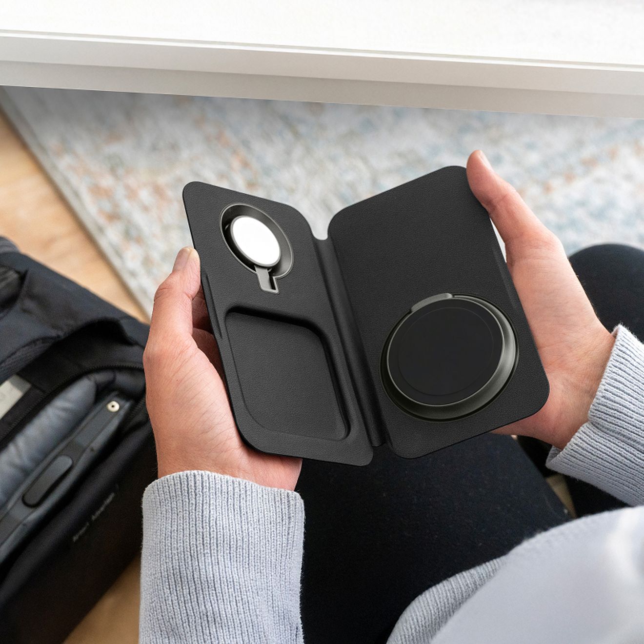 Una persona sostiene un cargador inalámbrico de viaje Nimble Fold 3 en 1 negro y desplegado, en el que se ven las zonas de carga específicas para un smartphone, un Apple Watch y unos audífonos inalámbricos.