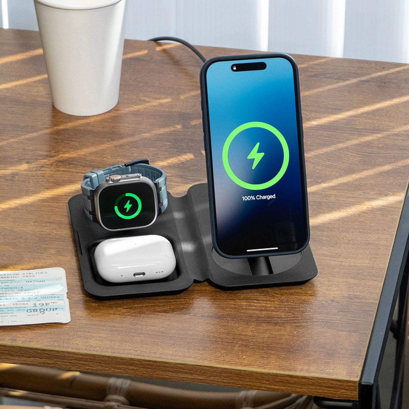 Un smartphone, un Apple Watch y un estuche para audífonos inalámbricos se cargan a la vez en un cargador inalámbrico de viaje Nimble Fold 3 en 1 negro, desplegado sobre una mesa de madera.