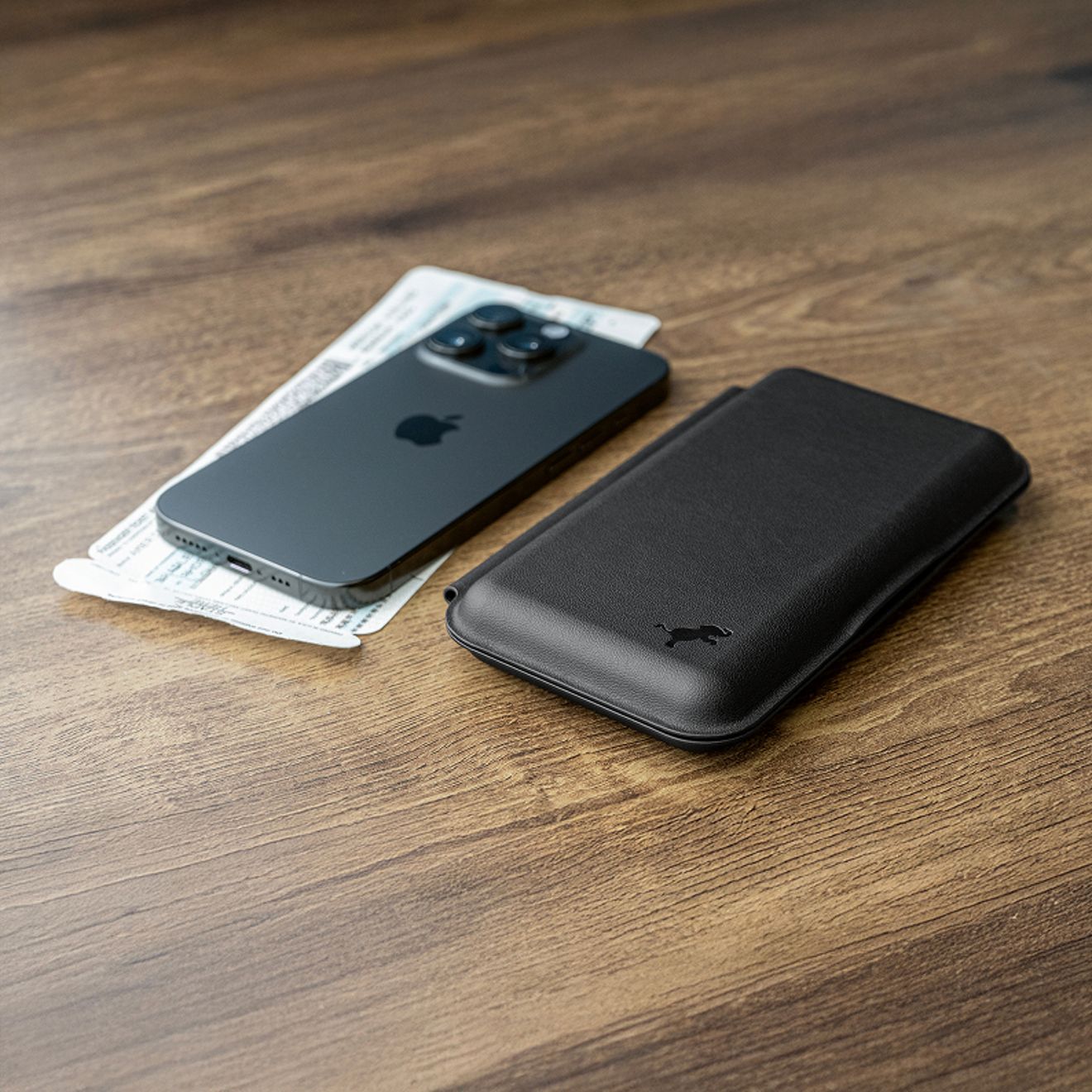 Un cargador inalámbrico Nimble Fold 3 en 1 negro, plegado junto a un iPhone y unas tarjetas de embarque sobre una mesa de madera.
