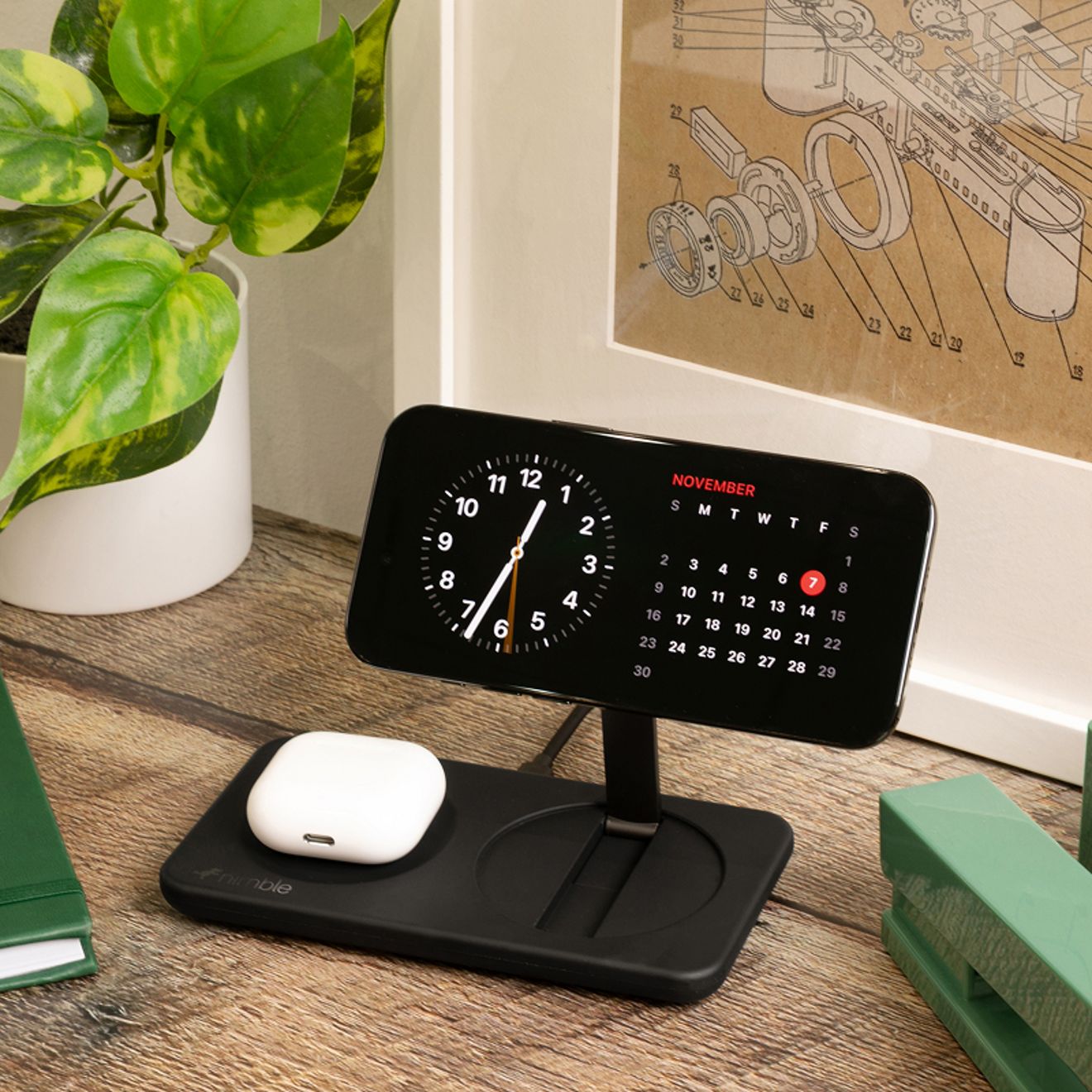 Un smartphone y un estuche de audífonos inalámbricos blancos se cargan simultáneamente en una base de carga inalámbrica magnética 2 en 1 Nimble de color negro sobre una mesita de noche de madera.