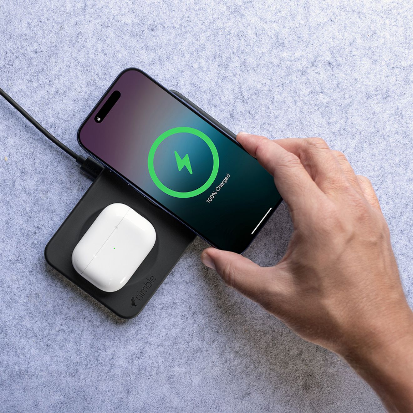 Una mano levanta un smartphone que muestra una carga completa, tomándolo de una base de carga inalámbrica 2 en 1 Nimble Podium en color negro, mientras un estuche blanco de audífonos inalámbricos se carga en la base.
