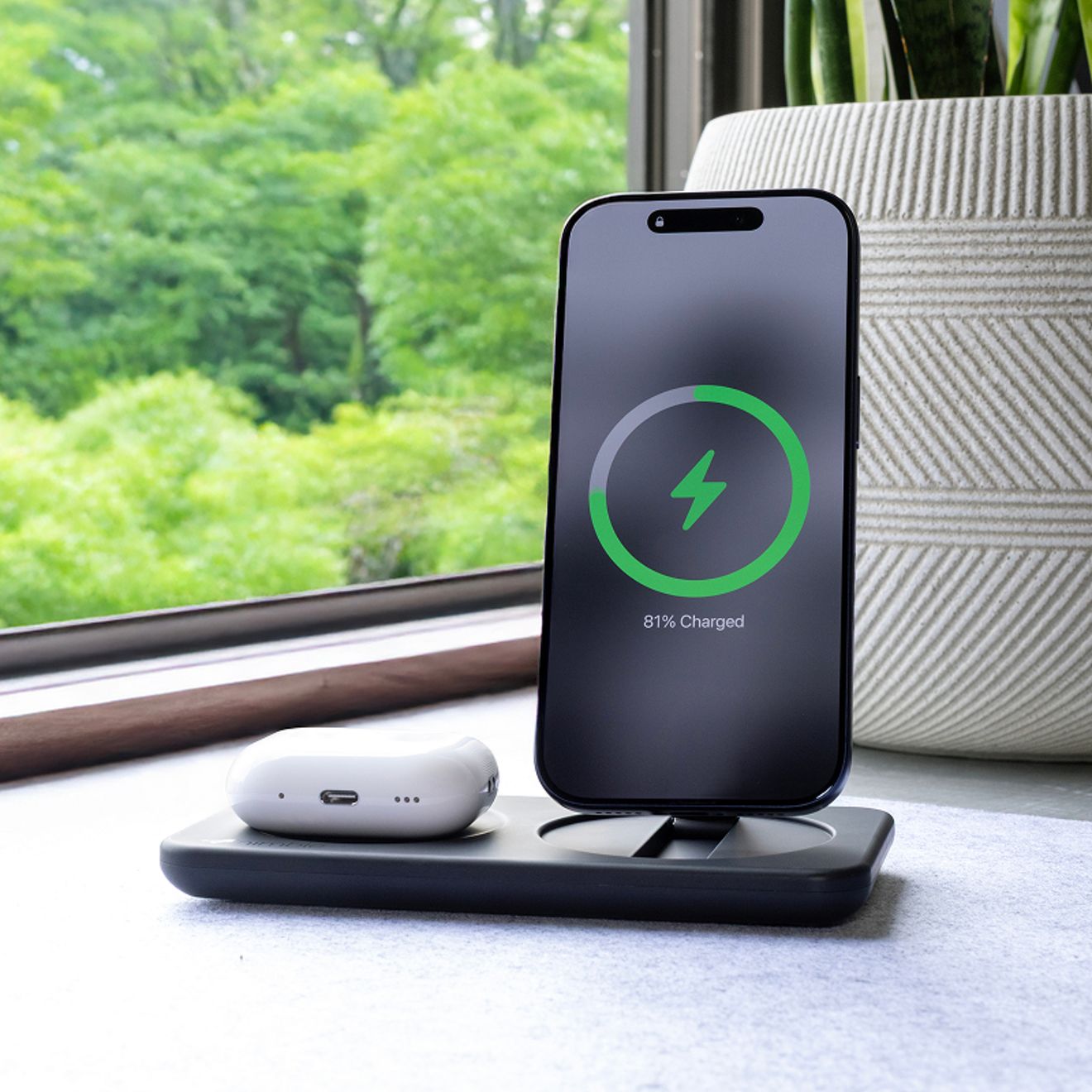 Un smartphone y un estuche blanco para audífonos inalámbricos se cargan simultáneamente en una base de carga inalámbrica Nimble negra, junto a una ventana.