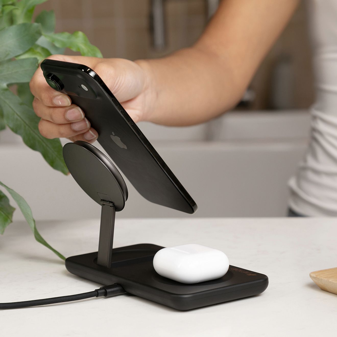 Una persona levanta un smartphone de una base de carga inalámbrica magnética 2 en 1 Nimble color negro, mientras un estuche blanco de audífonos inalámbricos se carga en la base.