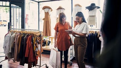 Una empleada en una tienda de ropa usando una tablet con POS para ayudar a una cliente a hacer una compra.
