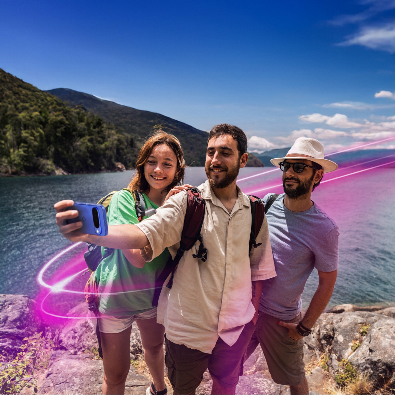 Tres amigos de pie en una colina rocosa rodeada de montañas y agua mientras se toman un selfie con un smartphone Pixel 10 y un aura magenta que los rodea.