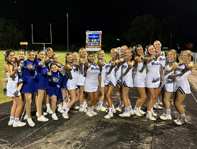 Las porristas de River Oaks, con uniformes blancos, posan para una foto en la pista del campo de fútbol.