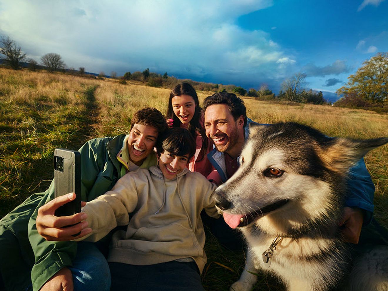 Clima otoñal al aire libre con un hombre, una mujer, una niña y un niño posando para un selfie con su perro, mostrando amistad y cercanía entre adultos y niños.