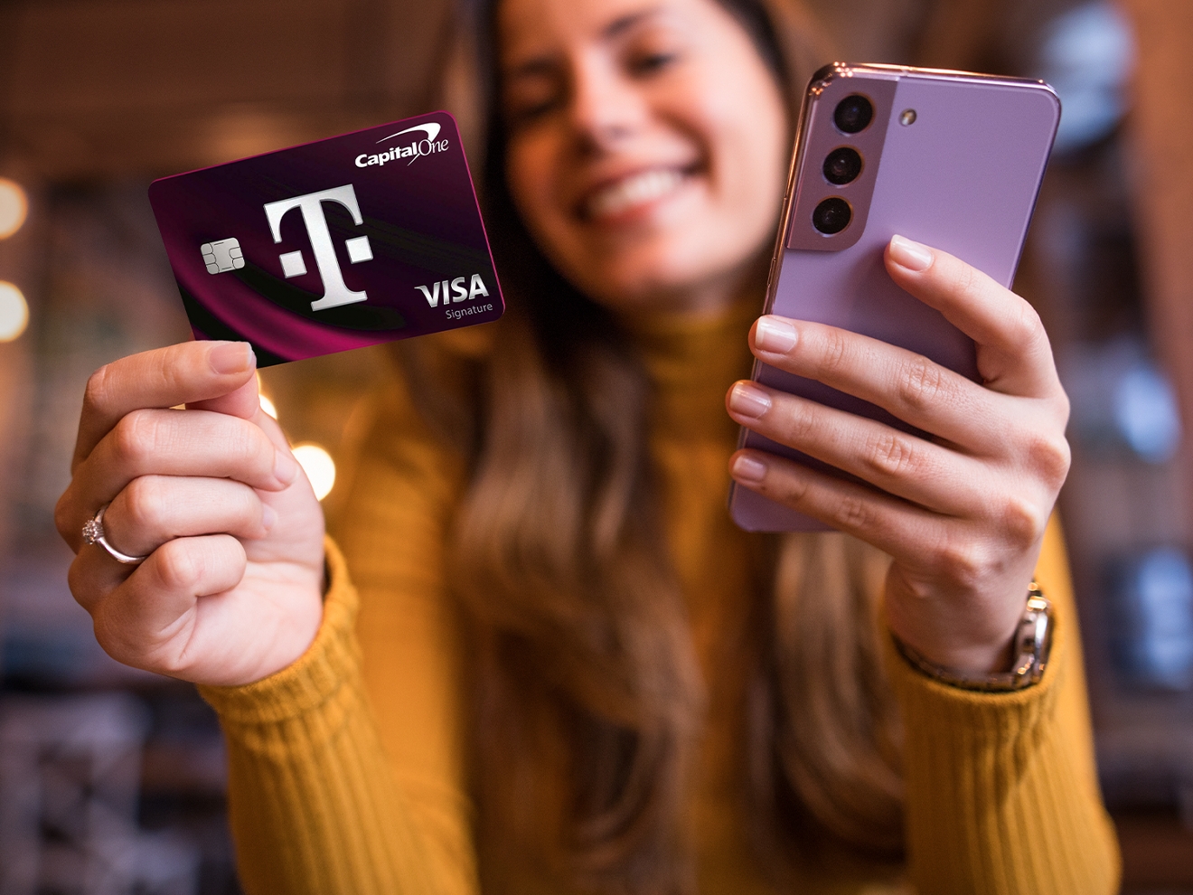 Una mujer sonríe mientras sostiene la tarjeta T-Mobile Visa en una mano y un teléfono en la otra.