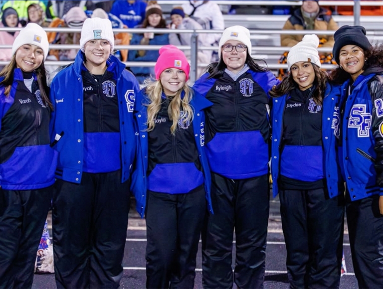 Las animadoras de Sanford-Fritch, vestidas con uniformes azules y negros a juego, posan para una foto en el borde del campo de fútbol.