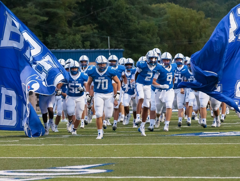 El equipo de fútbol americano de Brevard High School, con uniformes azules y blancos, entra al campo ondeando banderas.