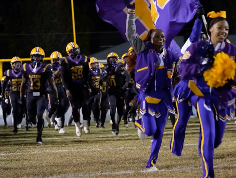 El equipo de fútbol de la Jackson High School y las animadoras, vestidas con uniformes morados y dorados, corren hacia el campo.