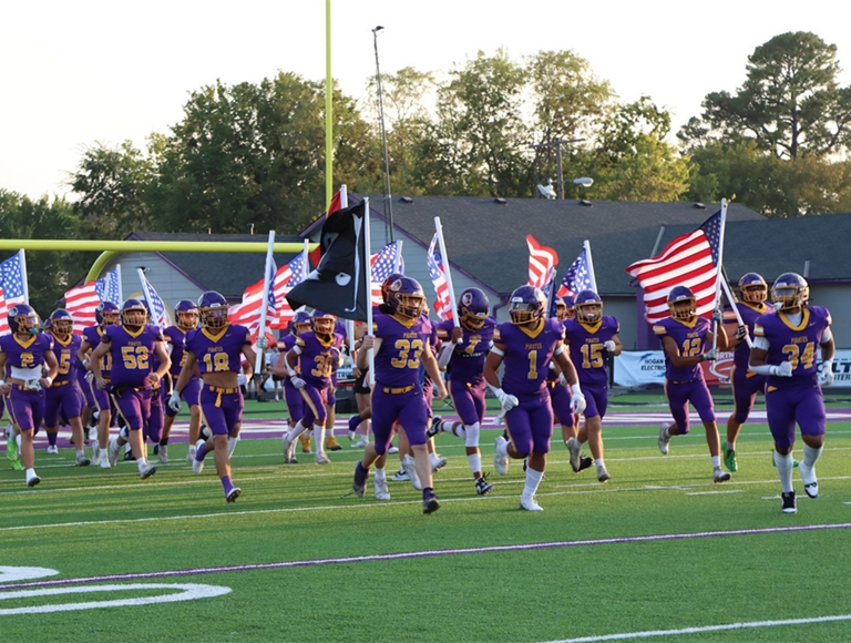 El equipo de fútbol americano de Bristow High School, con uniformes morados, entra al campo ondeando banderas.