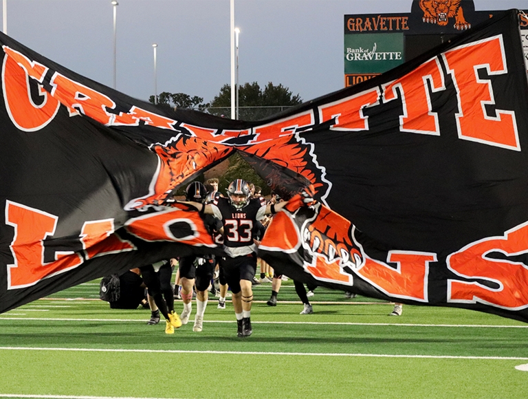 El equipo de fútbol americano de Gravette High School, vestido con uniformes naranjas y negros, corre a través de una pancarta gigante hacia el campo.
