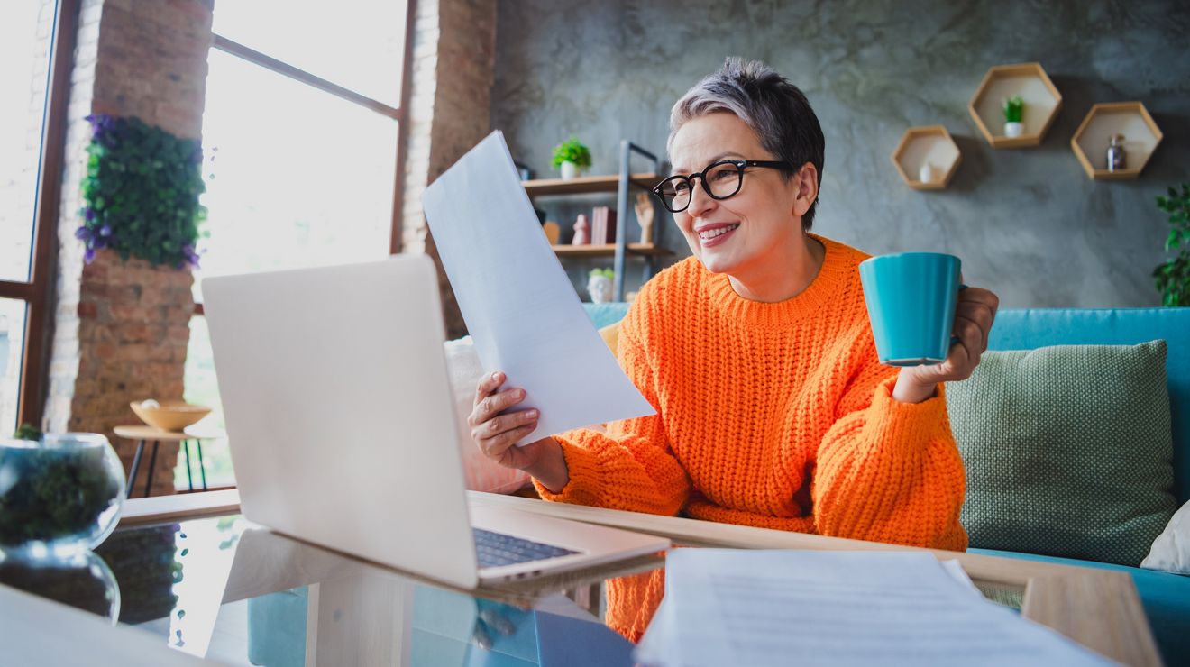 Foto de una mujer mayor sonriente y positiva con un suéter naranja trabajando con un dispositivo moderno y leyendo un informe en un apartamento; Shutterstock ID 2371415755; Client Name:Jeanette Delgado; Project #:State Relay; Project Name:State Relay; purchase_order:; job: