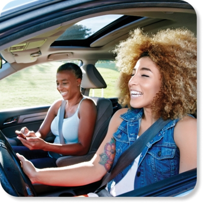 Una mujer sonriente conduce mientras otra mujer escribe en un teléfono.