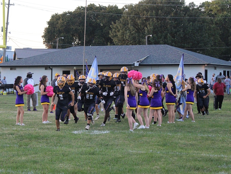Jugadores de fútbol americano de Slater High School corren hacia el campo pasando junto a las animadoras.