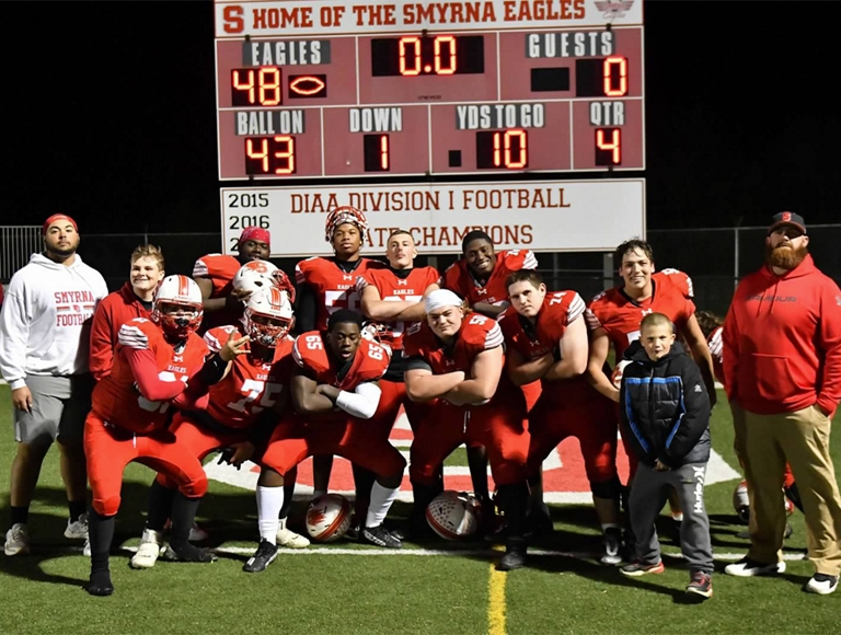El equipo de fútbol de Smyrna High School, con uniformes rojos, posa para una foto con un tablero de puntuación en el fondo.