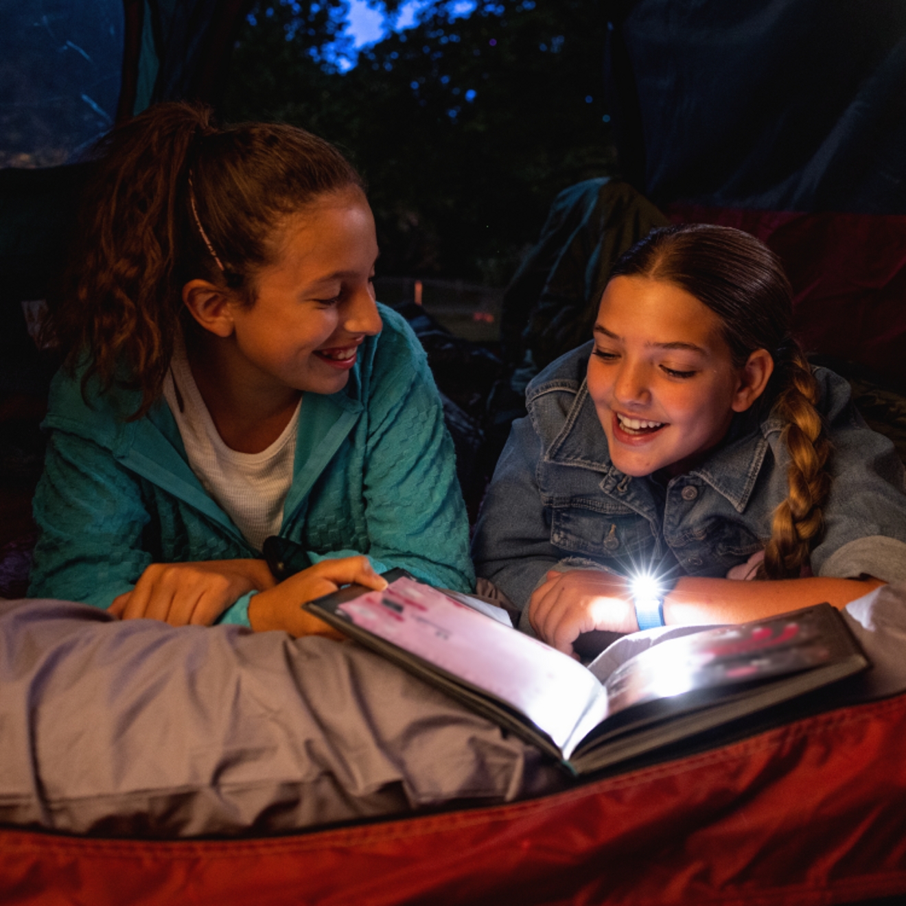 Dos niñas usando la linterna del reloj SyncUP KIDS para poder leer un libro en la oscuridad.