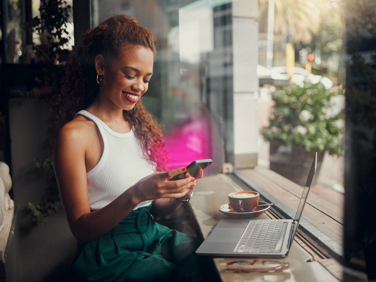 Una mujer usa su teléfono y su laptop mientras toma una taza de café. El teléfono emite un resplandor magenta de T-Mobile.