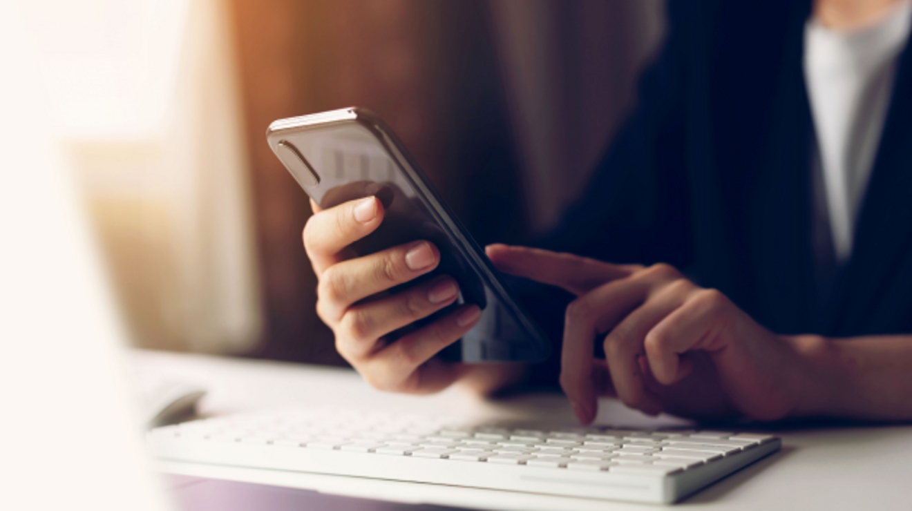 Primer plano de dos manos usando un smartphone sobre el teclado de una computadora.