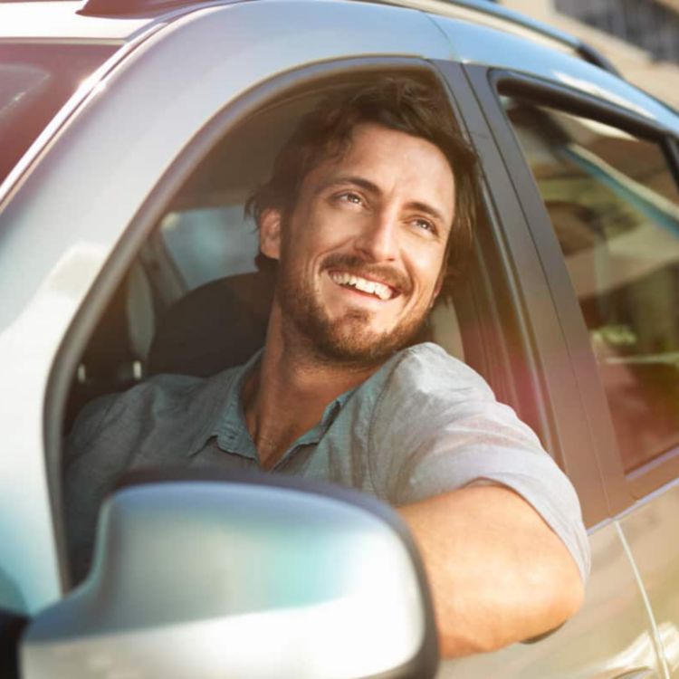 Un hombre sonriente mira por fuera de la ventanilla mientras conduce un auto alquilado.