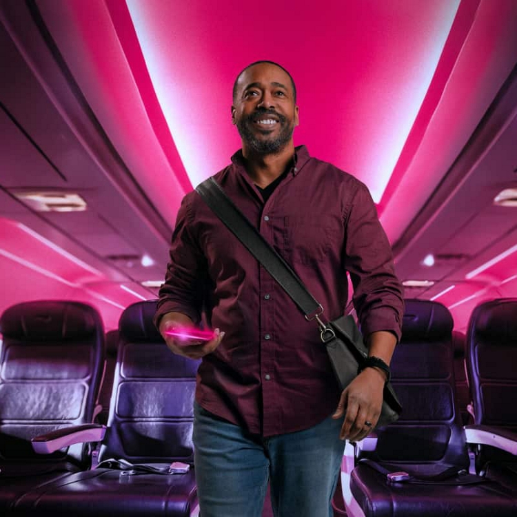 Un viajante de empresas sonriente camina por el pasillo del avión iluminado en magenta con el smartphone en la mano.