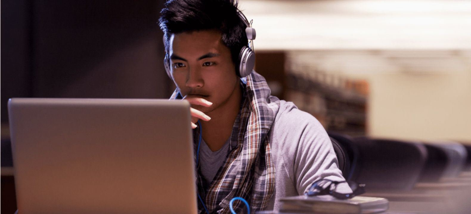 Un estudiante universitario con audífonos estudia en su laptop sentado en una biblioteca.