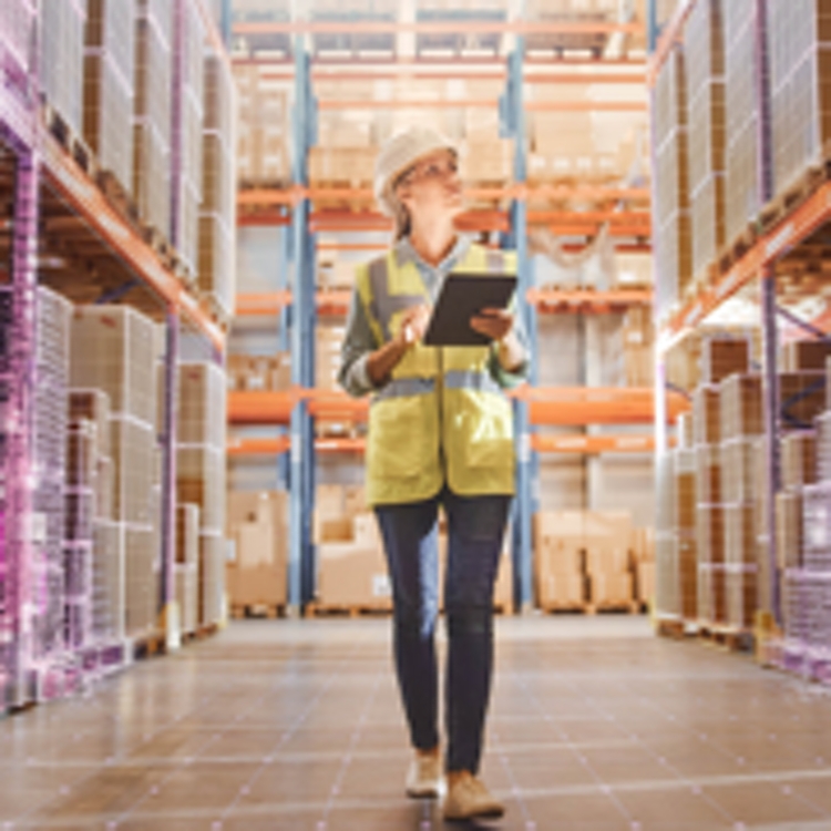 Mujer con chaleco amarillo lleva una tablet por el pasillo de un depósito con marcas magenta alrededor de las cajas en los estantes