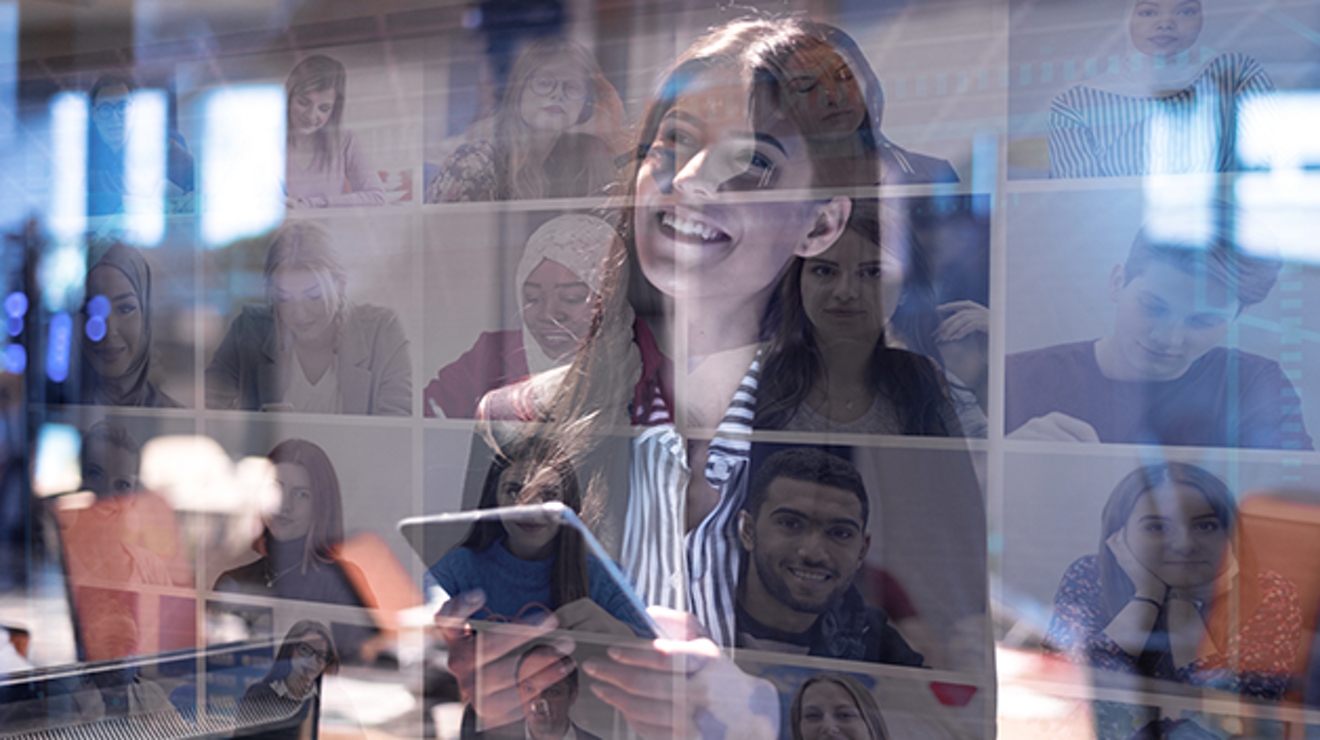 Mujer sonriente mirando una pantalla con otros participantes de una videoconferencia. Reflejo de ventanas en el fondo.