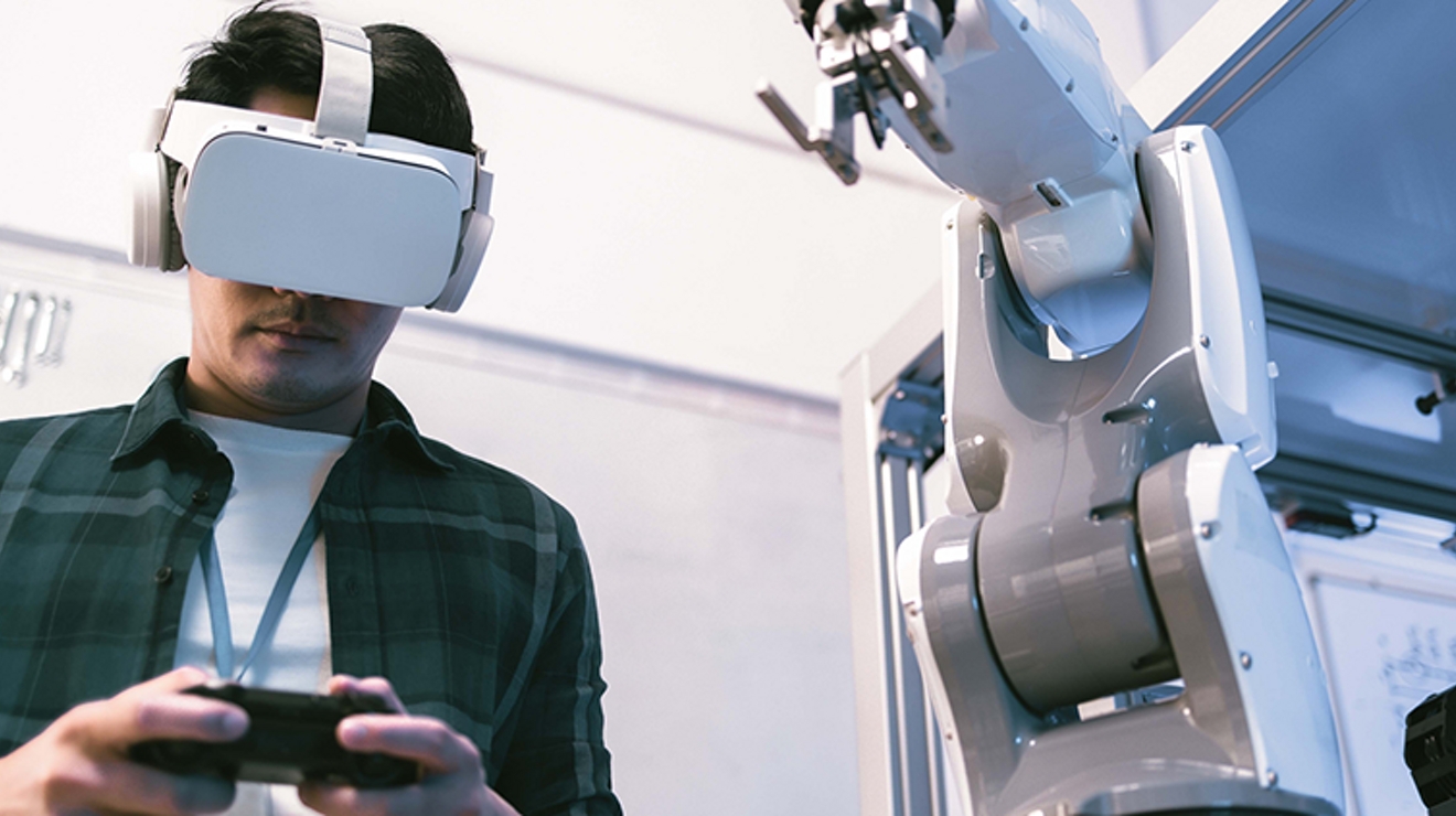 Un estudiante con un casco de RV de pie con las dos manos sosteniendo un dispositivo inteligente.
