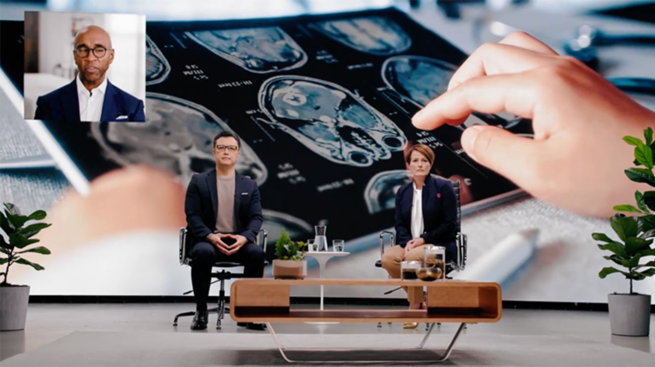 Dos personas sentadas en sillas en un escenario con una imagen gigante de una tablet que muestra resultados de una resonancia magnética.