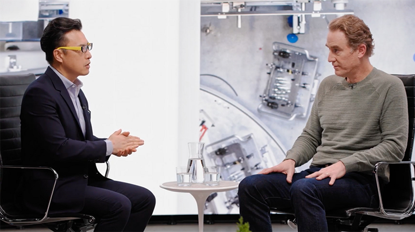 Dos hombres sentados en sillas en un escenario con una imagen gigante de dos personas reparando un brazo robotizado.