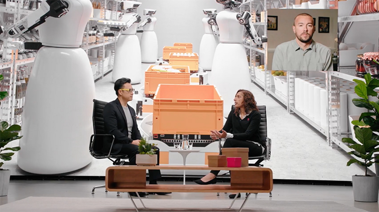 Dos personas sentados en sillas en un escenario con una imagen gigante de robots automatizados clasificando inventario.