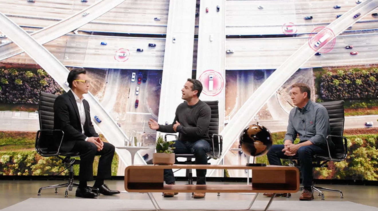 Dos hombres sentados en sillas en un escenario con una imagen gigante de una autopista en el fondo.